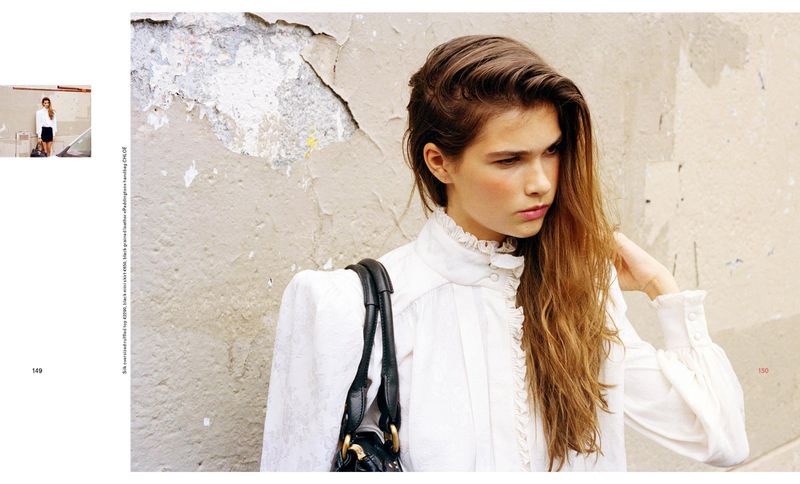 Brunette woman with long hair poses wearing a white ruffled shirt with a black leather purse in front of a textured wall.