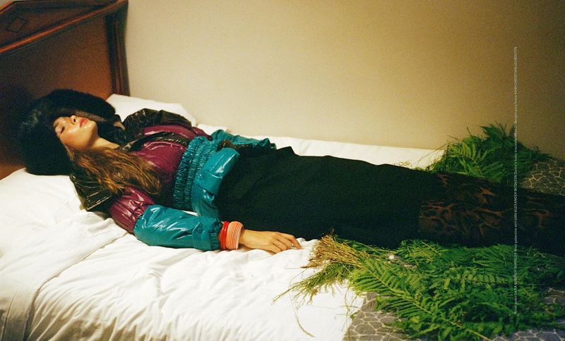 A woman with long hair lies on a bed in a purple, blue, and black jacket and black pants. Green plants are strewn on the bed.