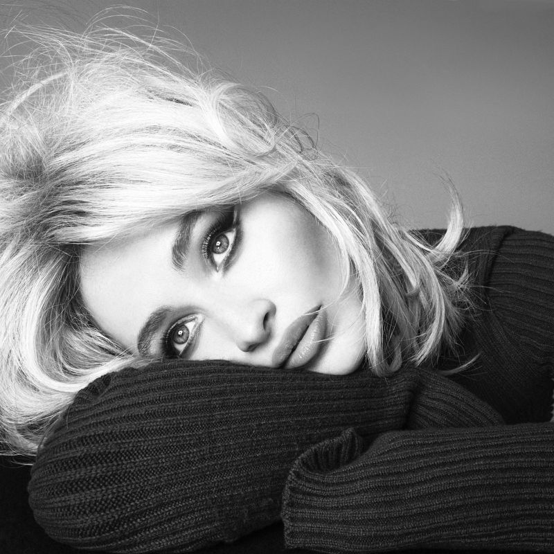 Monochrome portrait of a blond woman with makeup resting her head on her arms wearing a dark, knitted long-sleeve sweater against a gray backdrop.