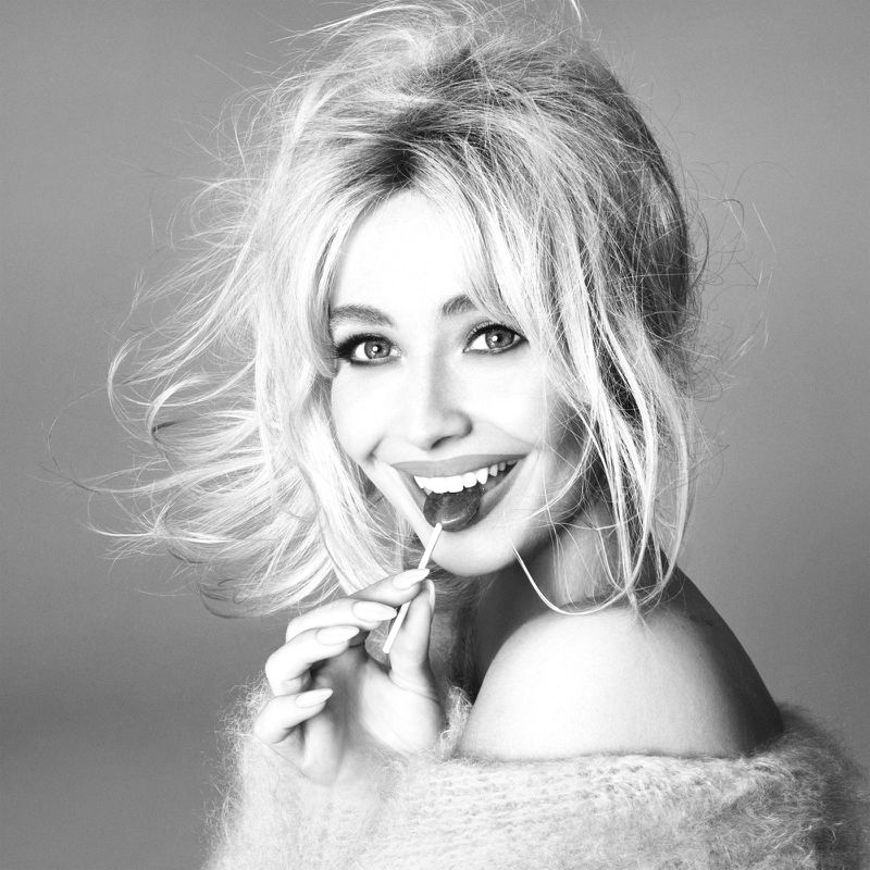 Black and white portrait of a smiling blonde woman with voluminous hair, wearing a fuzzy sweater and holding a lollipop to her mouth.