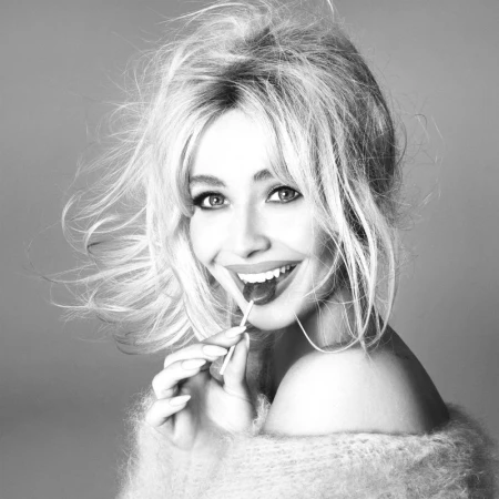 Black and white portrait of a smiling blonde woman with voluminous hair, wearing a fuzzy sweater and holding a lollipop to her mouth.