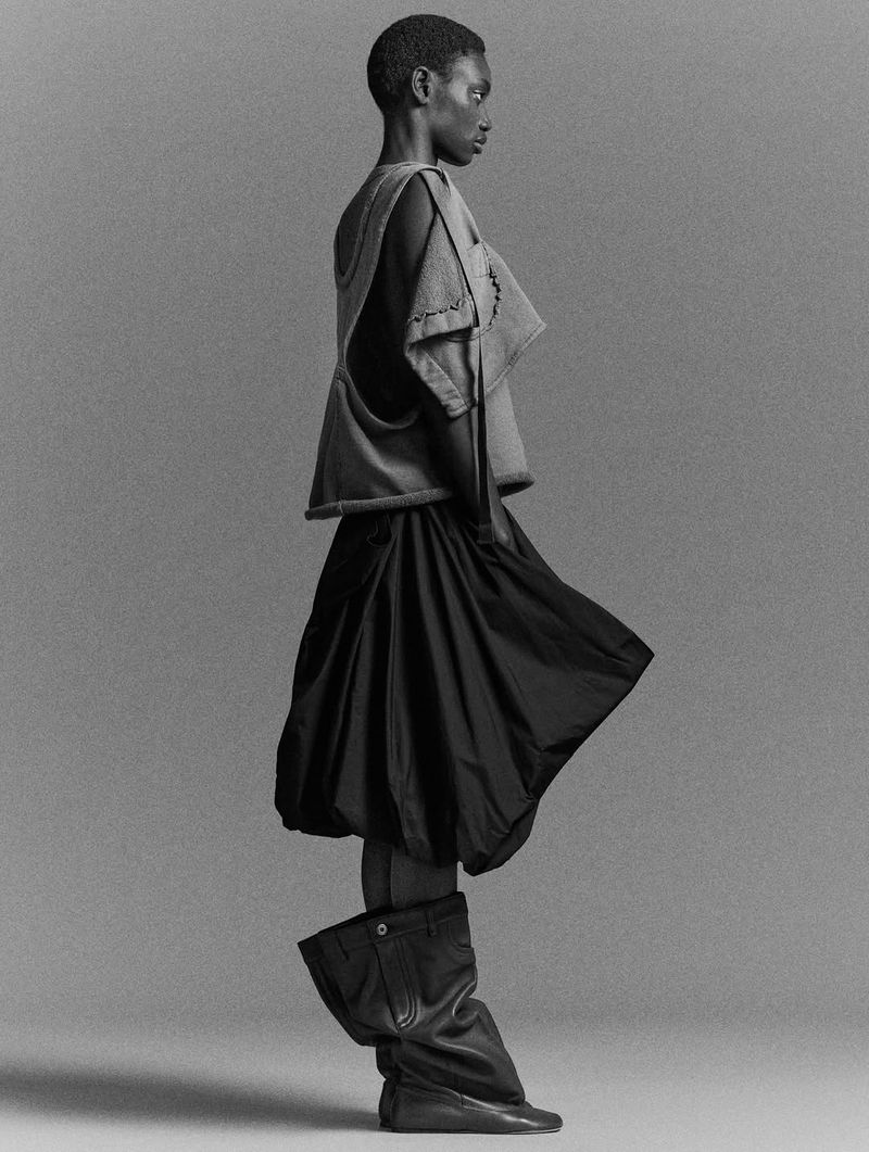 Monochrome fashion shot of a Black woman posing in a sleeveless top, voluminous skirt, and unique boots, against a plain grey background.