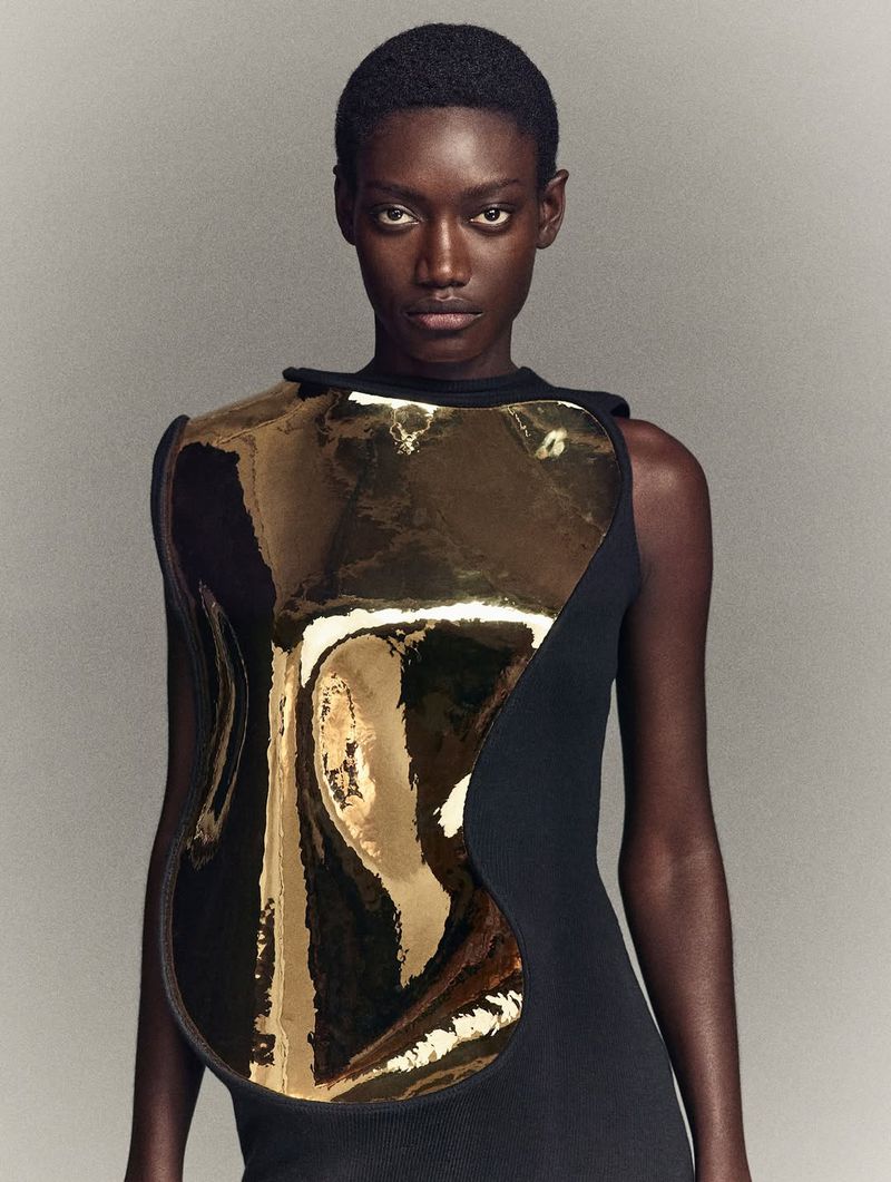 Striking studio portrait of a Black adult female model wearing a black dress with a gold metallic chest piece, gazing directly at the camera.