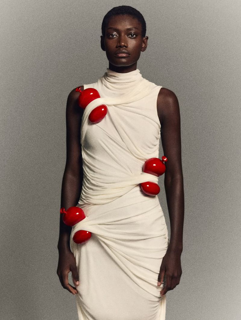 Fashionable woman with dark skin poses wearing a light-beige draped dress adorned with vibrant red, organic-shaped accents in a studio setting.
