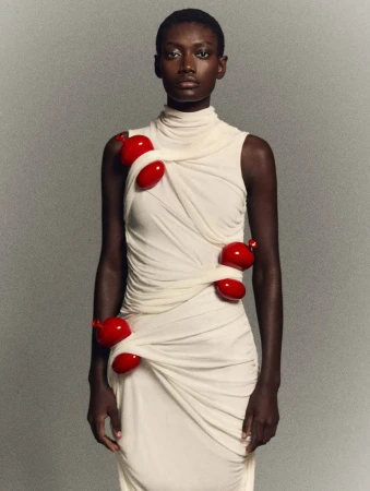 Fashionable woman with dark skin poses wearing a light-beige draped dress adorned with vibrant red, organic-shaped accents in a studio setting.