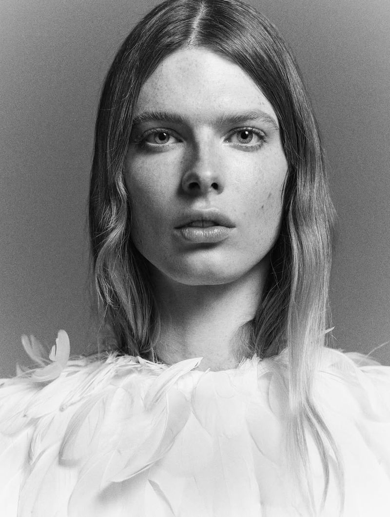 Monochrome portrait of a fair-skinned woman with long hair and feathers around her neck, set against a solid grey background.