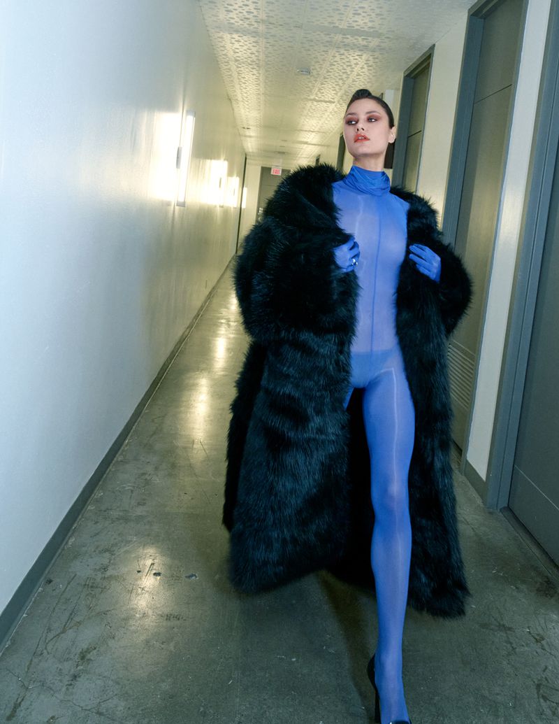 A woman in a blue bodysuit and black fur coat poses in a hallway with neutral walls and lighting, creating a modern fashion image.