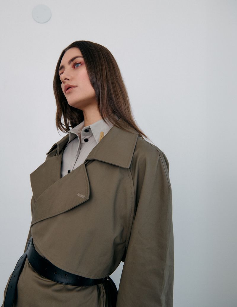 Portrait of a brunette woman wearing a trench coat and collared shirt against a white wall, looking off-camera in a studio setting.