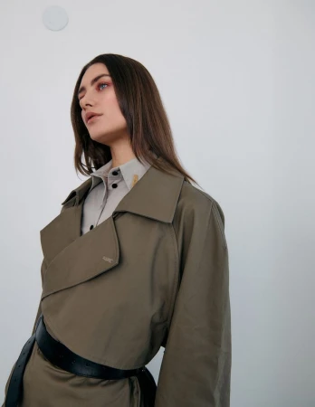 Portrait of a brunette woman wearing a trench coat and collared shirt against a white wall, looking off-camera in a studio setting.
