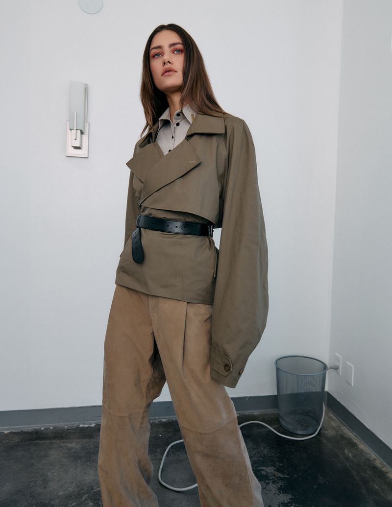 A woman in an olive green jacket and tan pants stands in a minimalist studio with white walls and concrete floors. She is wearing a black belt.