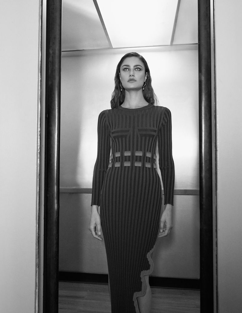 Black and white portrait of a slender woman wearing a long-sleeved ribbed dress with cutouts posing in front of a reflective background.
