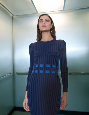 A woman with long hair in a long-sleeved blue dress poses in an elevator with silver walls and a grid-patterned ceiling with soft lighting.