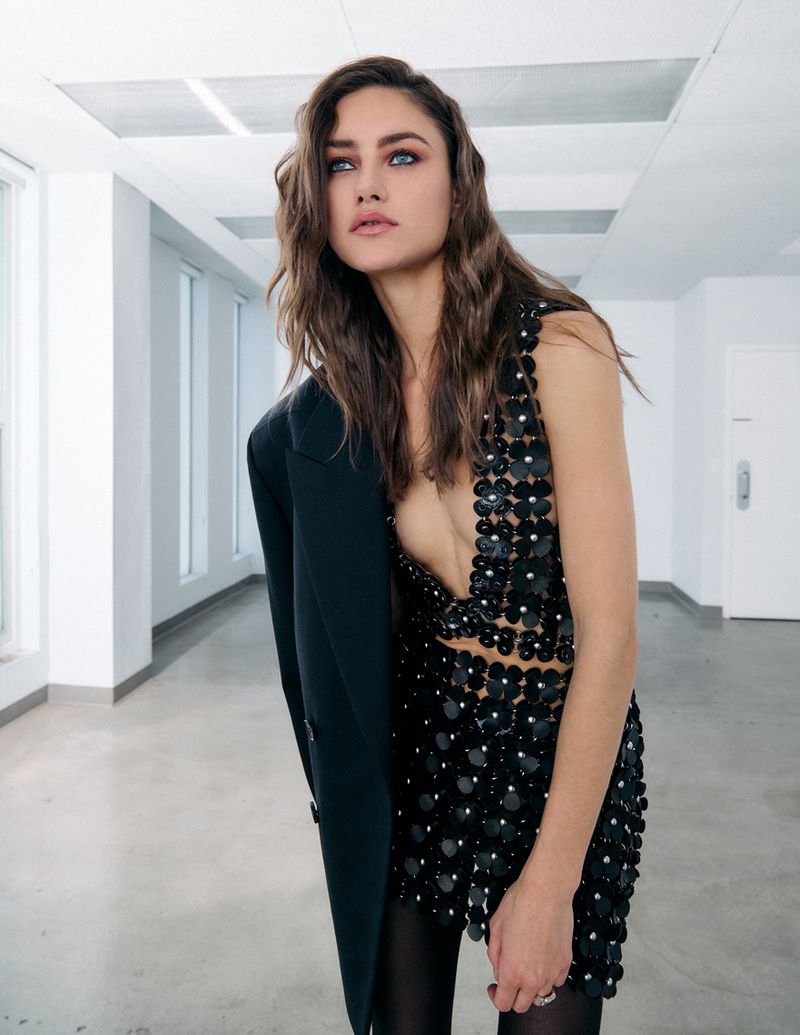 Attractive woman in a black, flower-sequined mini dress and black blazer poses indoors against a white wall with natural light.