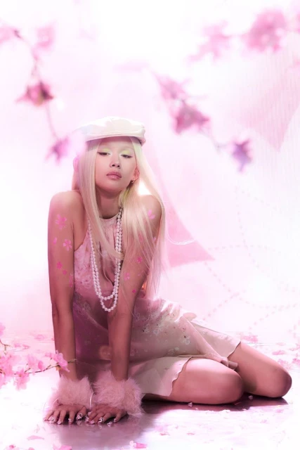 Young woman with platinum blonde hair poses with flowers, wearing pink dress, cap and pearl necklace in a soft, pink-toned photo studio.