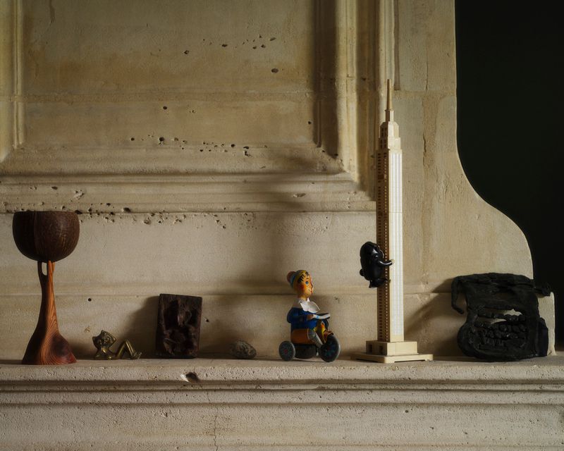Still life of assorted vintage figurines, including a toy climber on a miniature Empire State Building, arranged atop a stone mantelpiece.