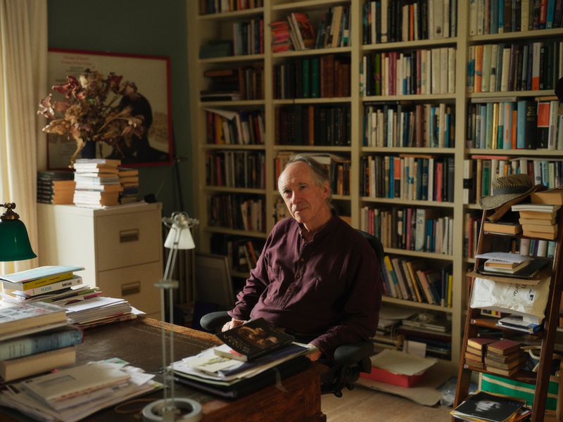 Elderly man sits in a chair surrounded by books. A desk with papers, filing cabinet, and a tall bookcase fill the room.