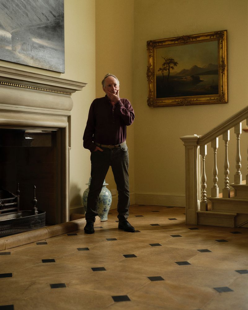 Mature man in brown shirt and grey pants stands in a home hallway with paintings, fireplace, staircase, and wooden floor.