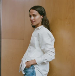 Alicia Vikander wearing a white button-down shirt and blue jeans against a wooden panel wall