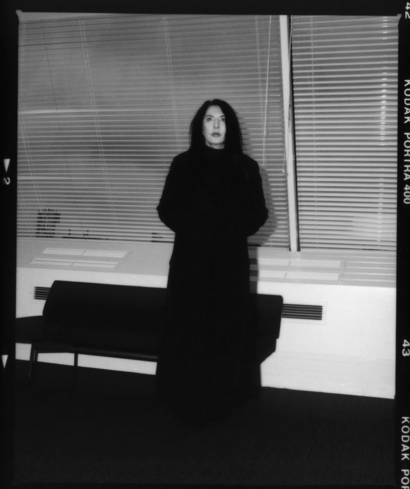 Full-length portrait of a woman in a long dark coat standing in an office space in front of a window with blinds. Black and white.