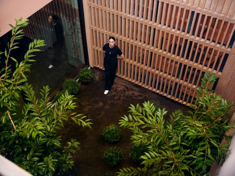Adult female in black uniform stands in a courtyard with green plants and a light-colored wooden-slat wall in the background.