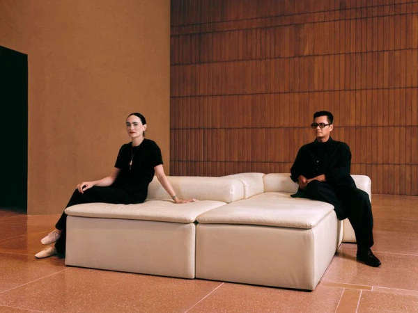 Two people seated on a modern white sectional sofa indoors with neutral tones and textures providing a minimalist architectural backdrop.