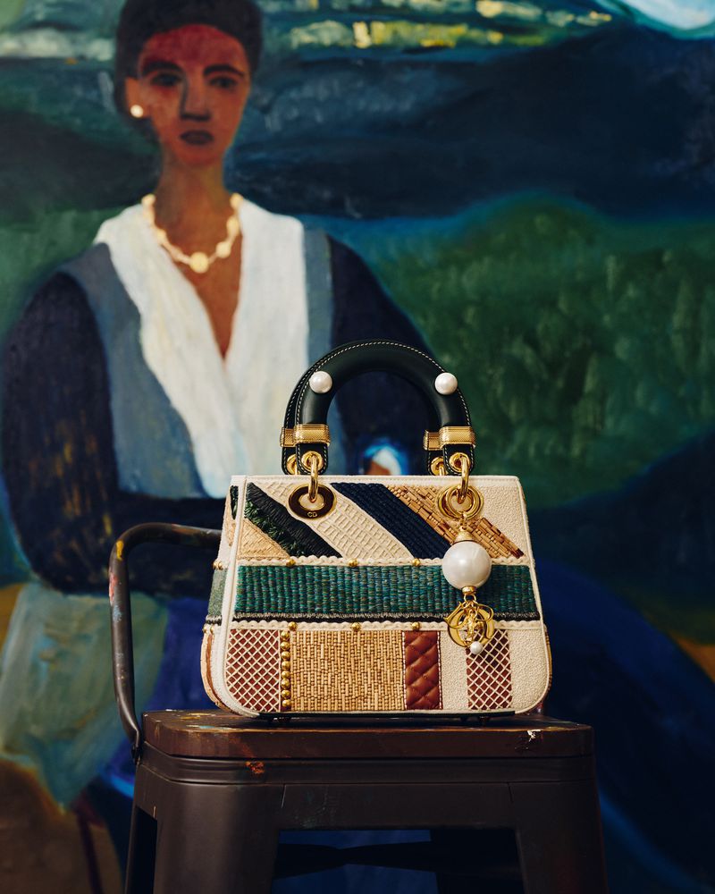 Lady Dior patchwork handbag displayed on a stool against an abstract painting in an indoor setting with studio lighting.