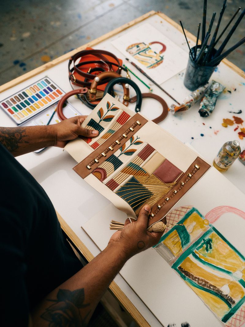 Person holding uniquely designed handbag with leather and fabric, displayed on a white table with art supplies and a concept sketch.