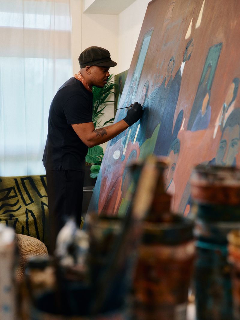 Artist at work in studio, painting large canvas with figures; natural light and painting supplies visible, wearing cap and black gloves.