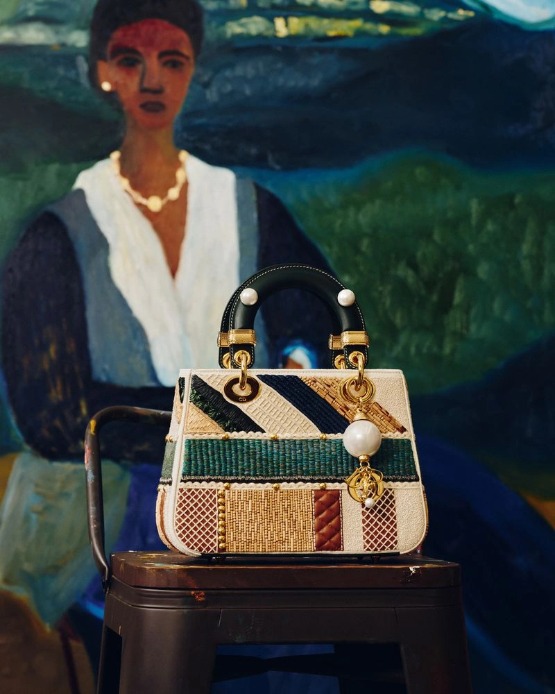 Dior Lady Art embroidered handbag resting on a dark stool in front of a painted figurative portrait background.