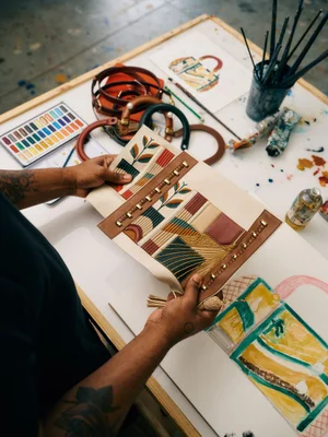 Artist holding an embroidered leather handbag panel over a studio worktable with sketches and paints.