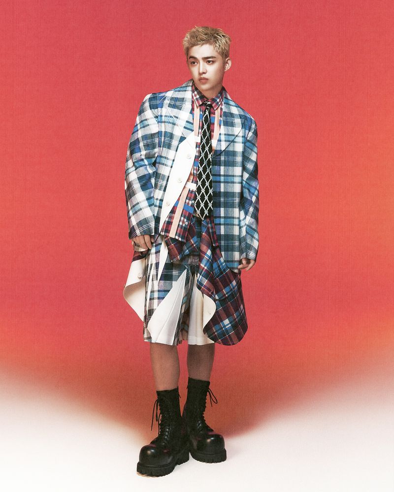 Full-length shot of a blonde male fashion model in a plaid outfit, tie and black platform boots against a gradient orange background.