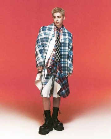 Full-length shot of a blonde male fashion model in a plaid outfit, tie and black platform boots against a gradient orange background.
