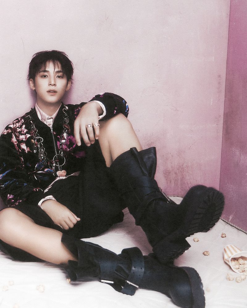 A stylish young adult sits on the floor in a floral jacket, dark pants, and boots. A pink background contrasts the dark outfit.