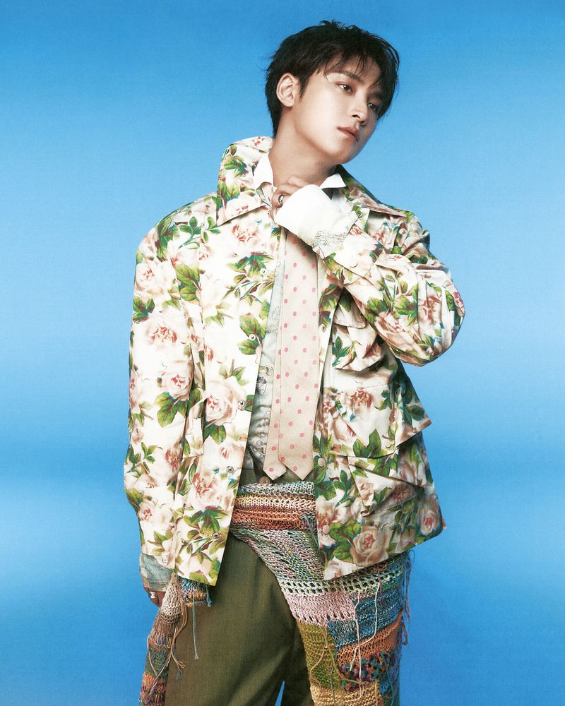 Studio shot of a handsome young man posing in floral print jacket, tie, and olive pants against a vibrant blue background.