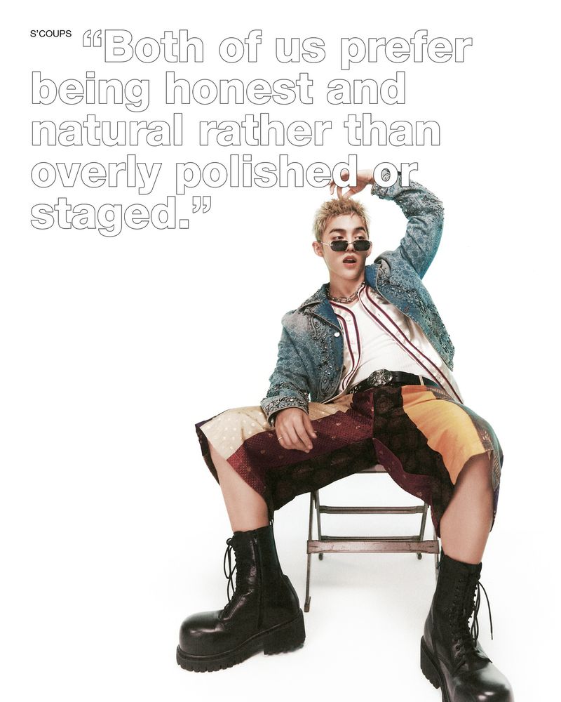 Stylish young adult posing with sunglasses indoors, wearing denim jacket, t-shirt, patchwork shorts and platform boots under bright lighting.