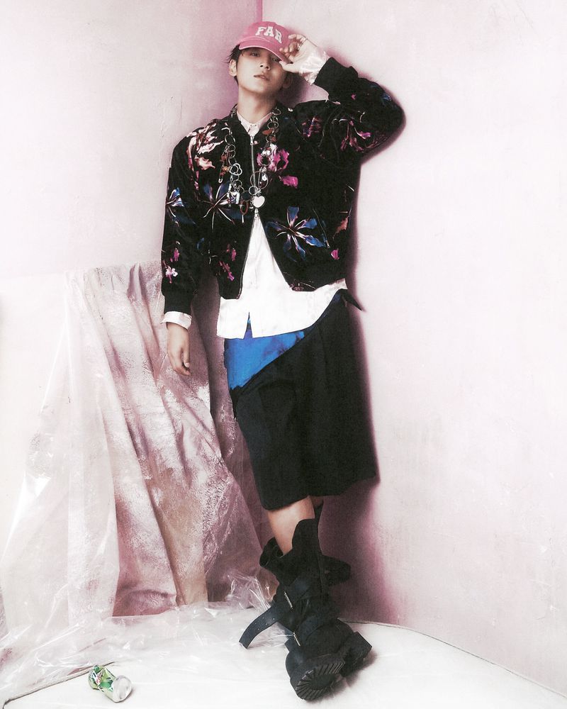 Young adult poses in a floral bomber jacket, shorts, and heavy boots against a light pink wall with draped plastic sheeting.
