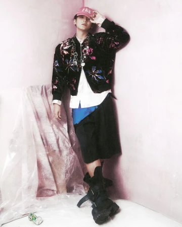 Young adult poses in a floral bomber jacket, shorts, and heavy boots against a light pink wall with draped plastic sheeting.