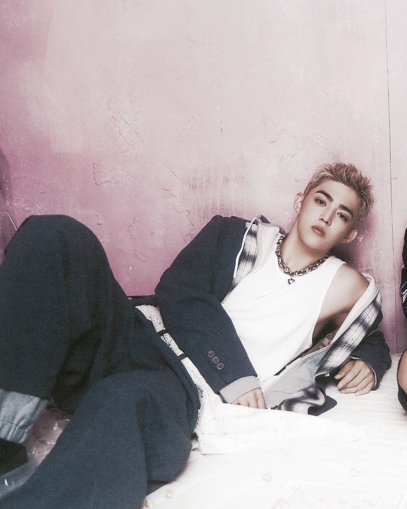 Young adult man poses while lying down indoors wearing a white tank top, a blazer jacket, and a chain necklace.