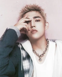 Young man with blonde spiky hair wearing a white t-shirt, plaid shirt, and dark jacket against a soft pink wall