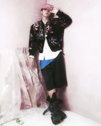 Young man wearing a floral bomber jacket, pink baseball cap, black shorts, and tactical boots leaning against a pink studio corner.