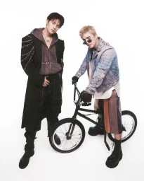 Two young men posing against a plain white background, one standing in a black coat and the other seated on a black bicycle wearing a patterned jacket.