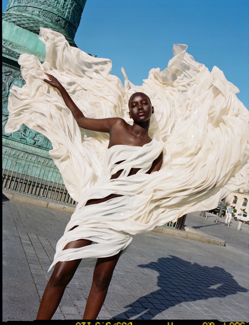 A fashionable African American woman poses outdoors in an elaborate flowing white dress with a blurred column and blue sky in the background.