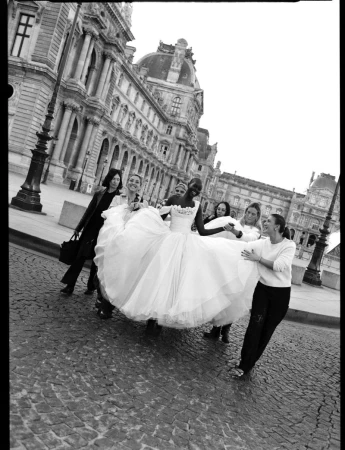 A bride in a white wedding dress is held up by a group of six people outdoors with historic architecture in the background in black and white.