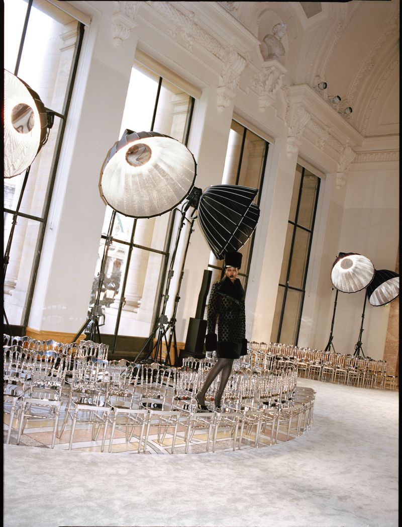 Fashion show scene: a runway featuring a model in black coat and hat surrounded by clear chairs and studio lighting equipment.