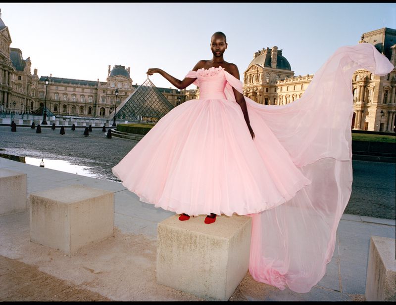 Fashion shot of a Black female model in a pink gown on a concrete block in front of the Louvre pyramid and museum in Paris.