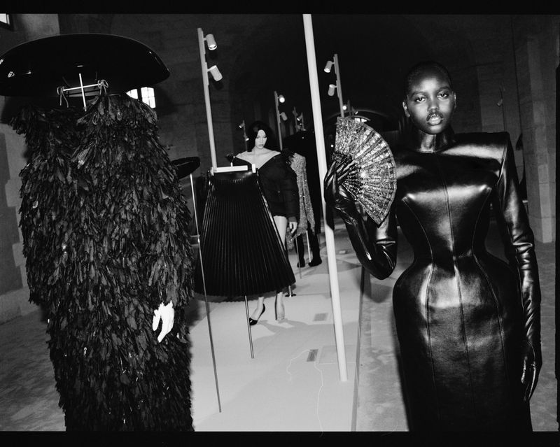 Runway fashion show featuring a Black model in fitted black dress holding fan, with mannequins in dark clothing in background, black and white.