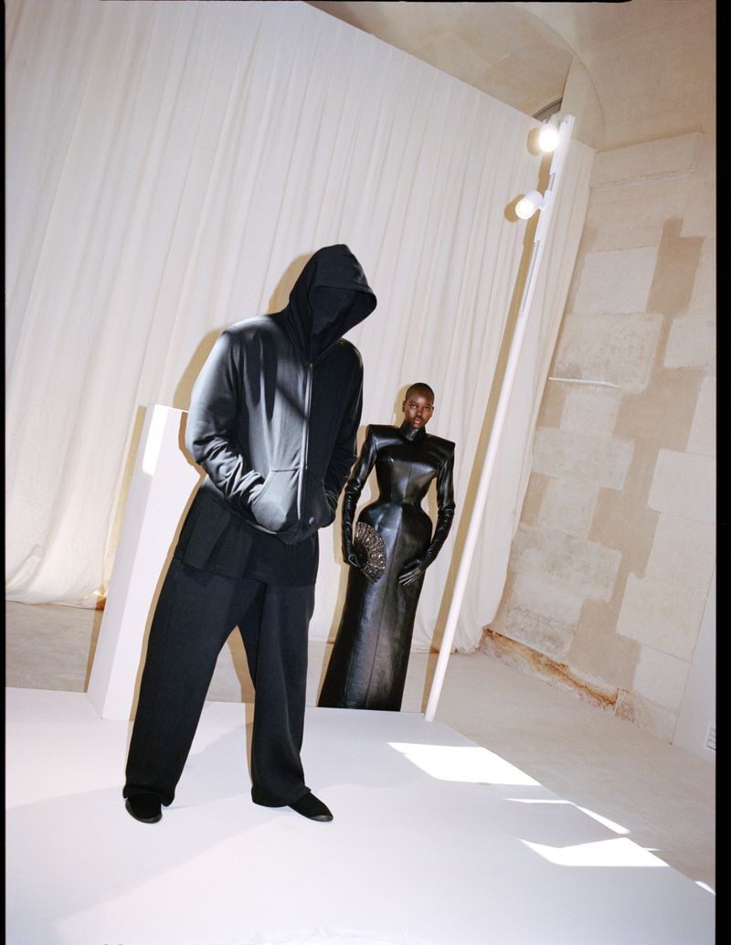 Two fashion models pose in all-black clothing against a bright neutral backdrop, one wearing a hooded jacket and the other a gown.