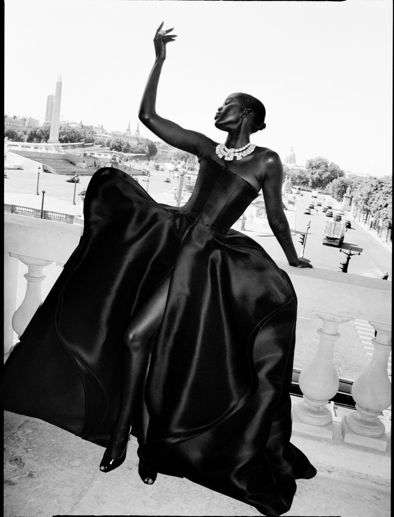 Elegant African woman in strapless black dress poses on balcony with city view, featuring monuments and traffic in black and white.