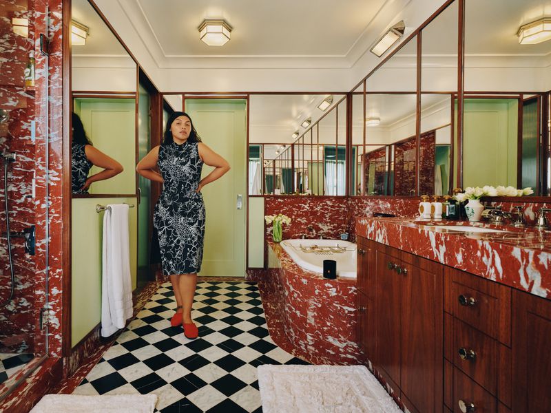 Woman in patterned dress and red flats poses in a bathroom with black and white checkered floor, marble walls, and vintage fixtures.