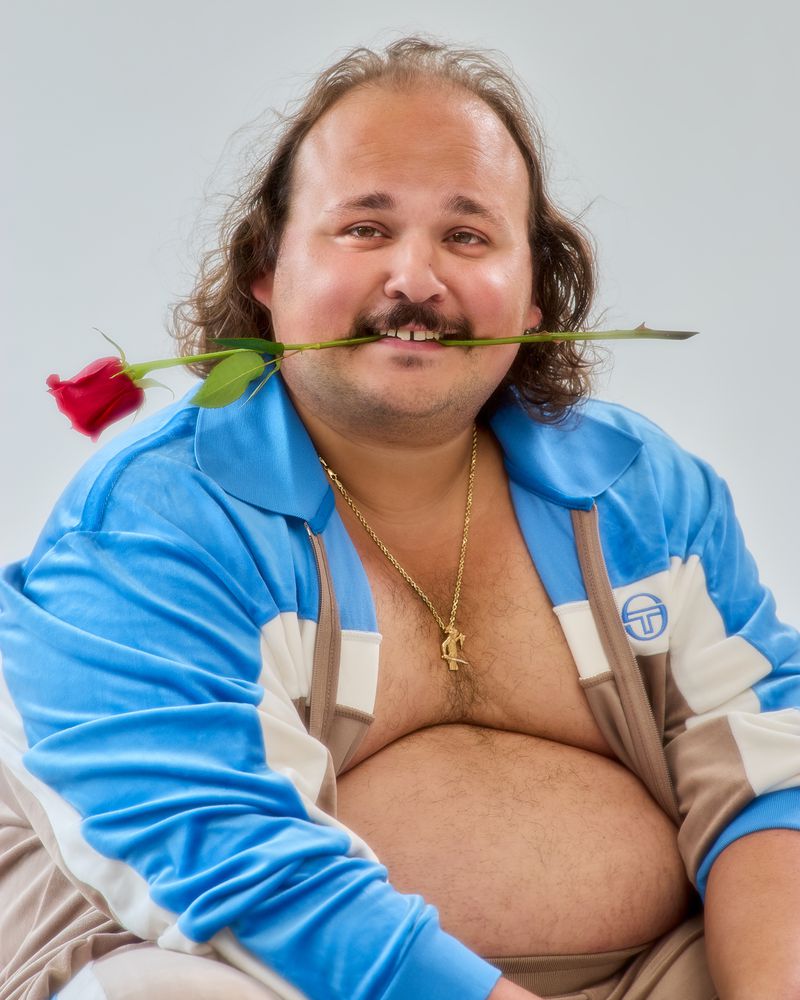 Man with mustache holds a red rose in his mouth, wearing blue-and-white tracksuit with gold necklace on plain white background.
